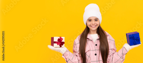 happy child in hat and mittens choose present box on yellow background, choice. Banner of christmas child girl, studio kid winter portrait with copy space.
