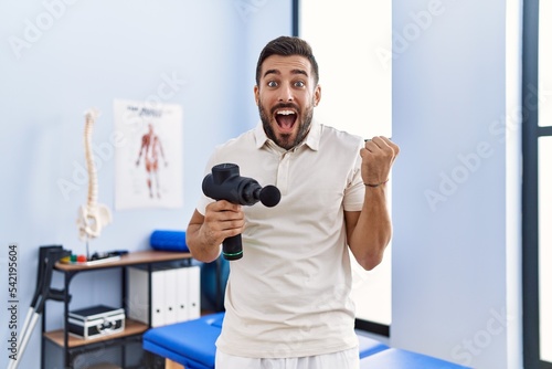 Handsome hispanic man holding therapy massage gun at physiotherapy center screaming proud, celebrating victory and success very excited with raised arms