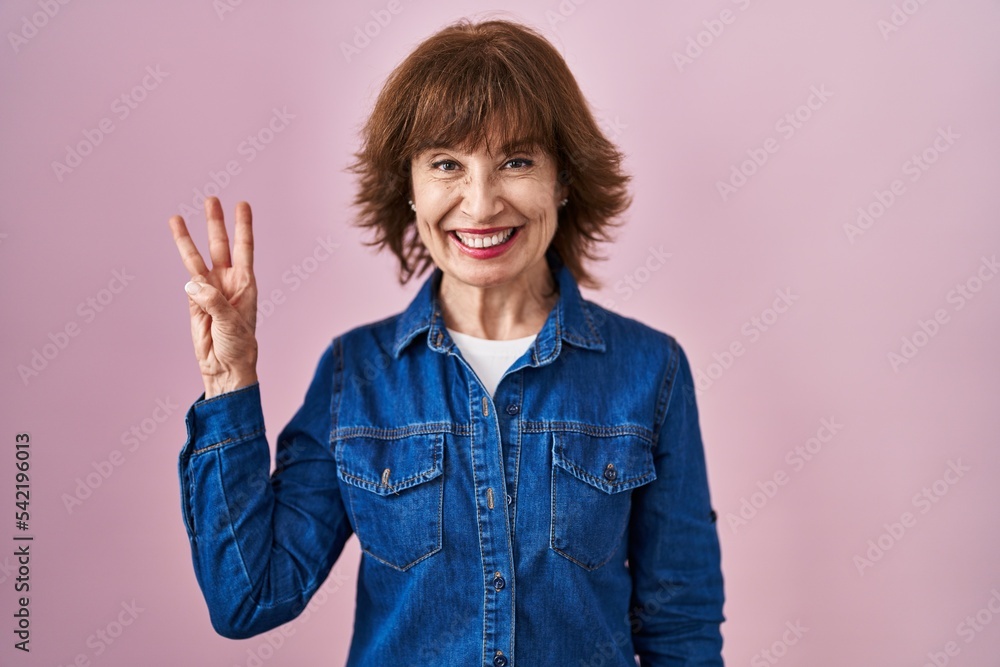 Middle age woman standing over pink background showing and pointing up with fingers number three while smiling confident and happy. Middle age woman standing over pink background showing and pointing up with fingers number three while smiling confident and happy.