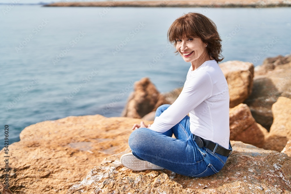 Middle age woman smiling confident sitting on the rock at seaside