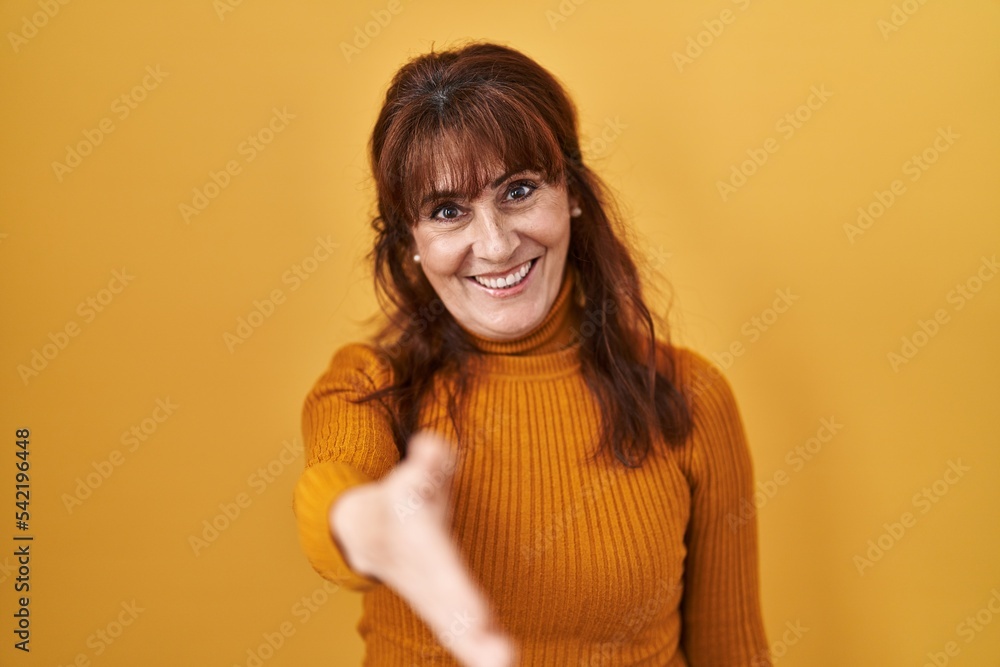 Middle age hispanic woman standing over yellow background smiling friendly offering handshake as greeting and welcoming. successful business. Middle age hispanic woman standing over yellow background smiling friendly offering handshake as greeting and welcoming. successful business.