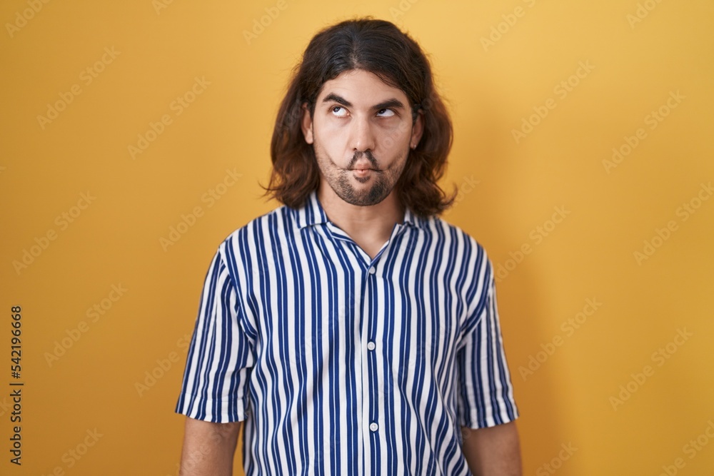 Hispanic man with long hair standing over yellow background making fish face with lips, crazy and comical gesture. funny expression. Hispanic man with long hair standing over yellow background making fish face with lips, crazy and comical gesture. funny expression.
