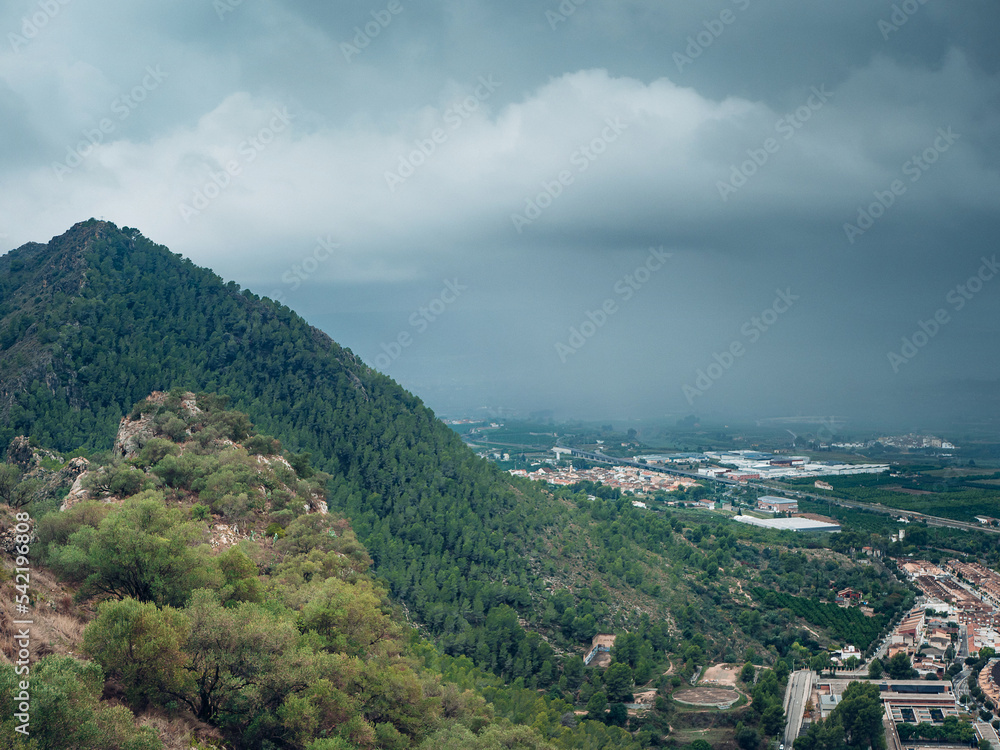 A small town under a mountain to which heavy rain and storm clouds is moving