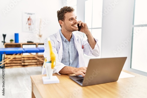 Young hispanic man wearing physiotherapist uniform using laptop talking on the smartphone at clinic