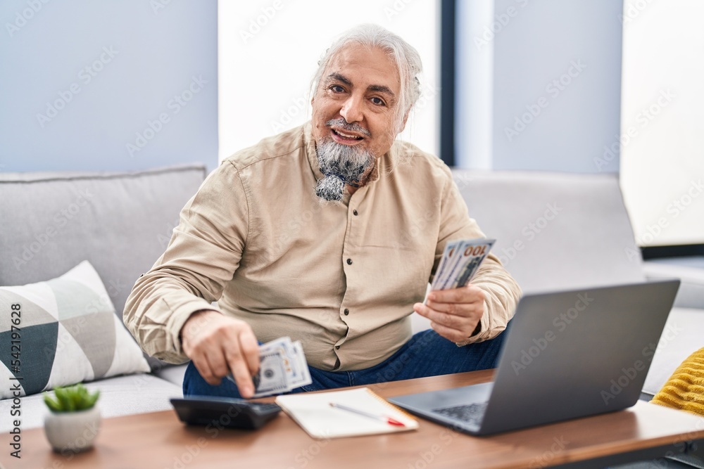 Middle age grey-haired man using laptop counting dollars at home Middle age grey-haired man using laptop counting dollars at home