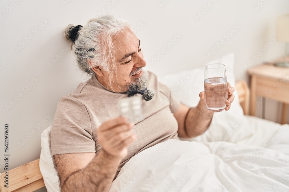 Middle age grey-haired man holding pills tablet and glass of water sitting on bed at bedroom Middle age grey-haired man holding pills tablet and glass of water sitting on bed at bedroom