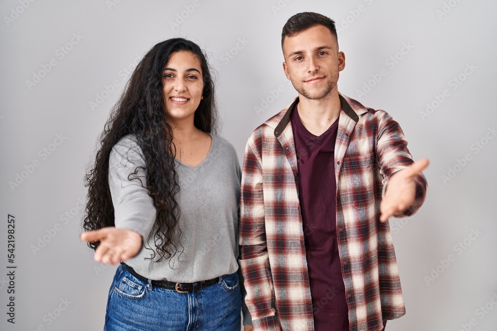 Young hispanic couple standing over white background smiling cheerful offering palm hand giving assistance and acceptance. Young hispanic couple standing over white background smiling cheerful offering palm hand giving assistance and acceptance.