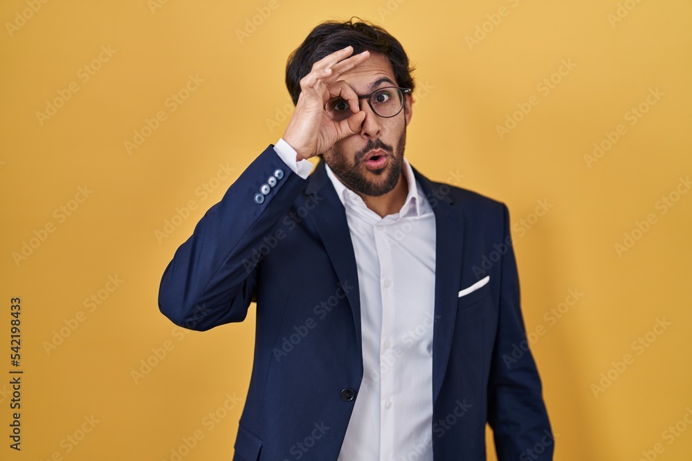 Handsome latin man standing over yellow background doing ok gesture shocked with surprised face, eye looking through fingers. unbelieving expression. Handsome latin man standing over yellow background doing ok gesture shocked with surprised face, eye looking through fingers. unbelieving expression.