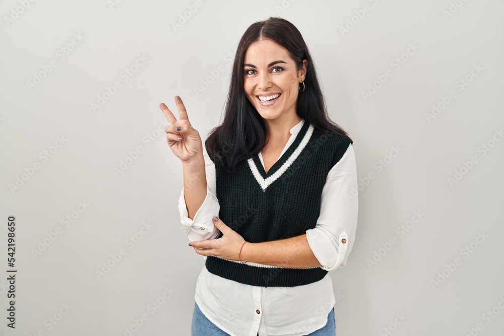 Young hispanic woman standing over isolated background smiling with happy face winking at the camera doing victory sign. number two. Young hispanic woman standing over isolated background smiling with happy face winking at the camera doing victory sign. number two.