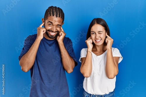 Young hispanic couple standing together covering ears with fingers with annoyed expression for the noise of loud music. deaf concept.