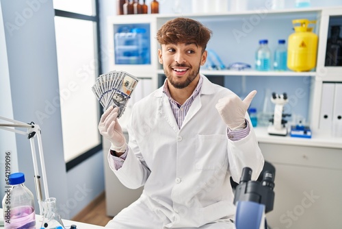 Arab man with beard working at scientist laboratory holding money pointing thumb up to the side smiling happy with open mouth