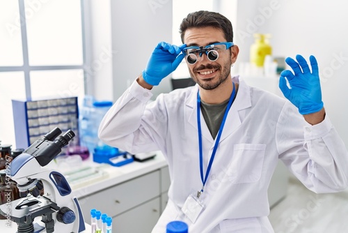 Young hispanic man working at scientist laboratory wearing magnifying glasses doing ok sign with fingers  smiling friendly gesturing excellent symbol