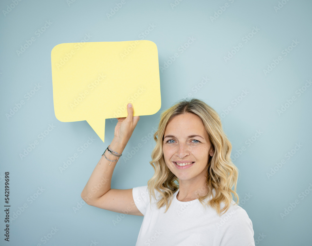pretty young woman with blond curls and white t-shirt is standing in front of blue wall and holding speech bubble pretty young woman with blond curls and white t-shirt is standing in front of blue wall and holding speech bubble