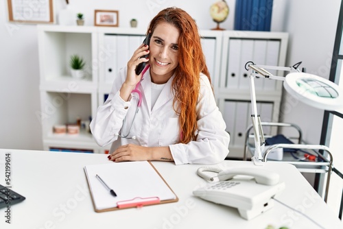Young redhead woman wearing doctor uniform talking on the smartphone at hospital