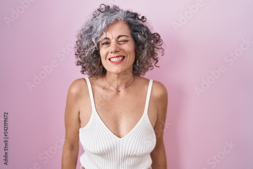 Middle age woman with grey hair standing over pink background winking looking at the camera with sexy expression, cheerful and happy face.