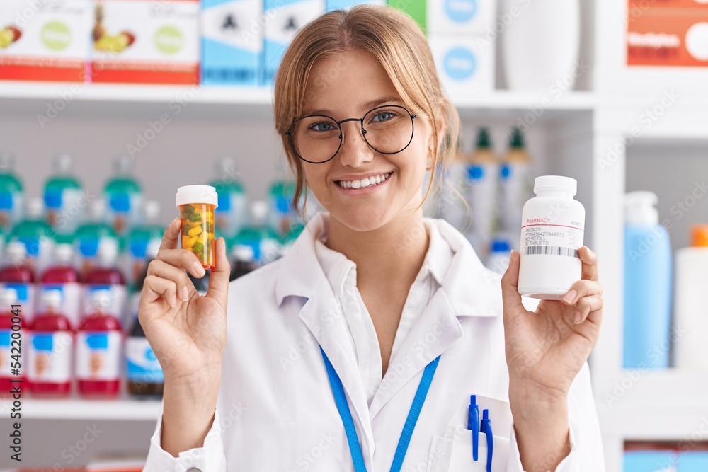 Young blonde girl pharmacist smiling confident holding pills bottles at pharmacy