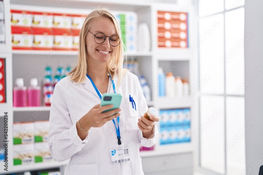 Young blonde woman pharmacist holding pills bottle using smartphone at pharmacy