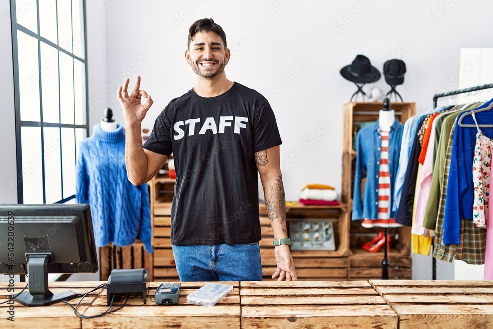 Young hispanic man working at retail boutique smiling positive doing ok sign with hand and fingers. successful expression.
