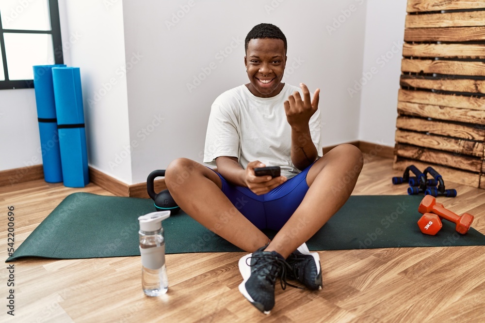 Young african man sitting on training mat at the gym using smartphone beckoning come here gesture with hand inviting welcoming happy and smiling Young african man sitting on training mat at the gym using smartphone beckoning come here gesture with hand inviting welcoming happy and smiling