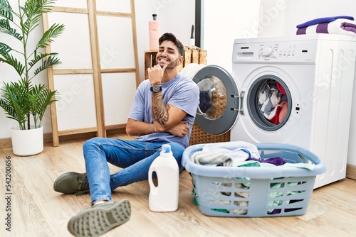 Young hispanic man putting dirty laundry into washing machine with hand on chin thinking about question, pensive expression. smiling and thoughtful face. doubt concept.
