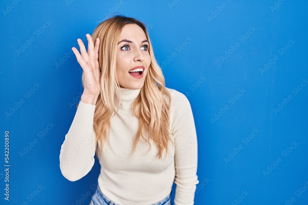 Young caucasian woman standing over blue background smiling with hand over ear listening an hearing to rumor or gossip. deafness concept.