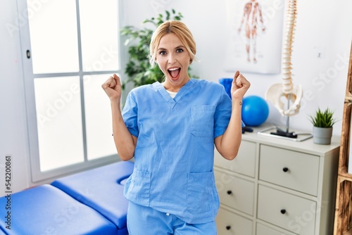 Beautiful blonde physiotherapist woman working at pain recovery clinic celebrating surprised and amazed for success with arms raised and open eyes. winner concept.