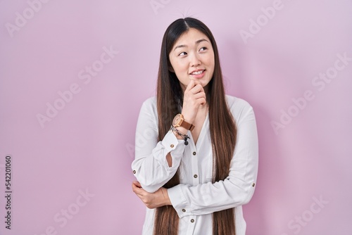 Chinese young woman standing over pink background with hand on chin thinking about question, pensive expression. smiling and thoughtful face. doubt concept.