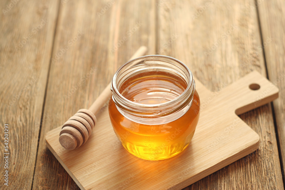 A transparent jar of honey stands on a wooden table. Nearby lies a honey stick A transparent jar of honey stands on a wooden table. Nearby lies a honey stick