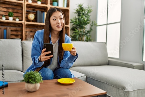 Young chinese girl using smartphone and drinking coffee sitting on the sofa at home.