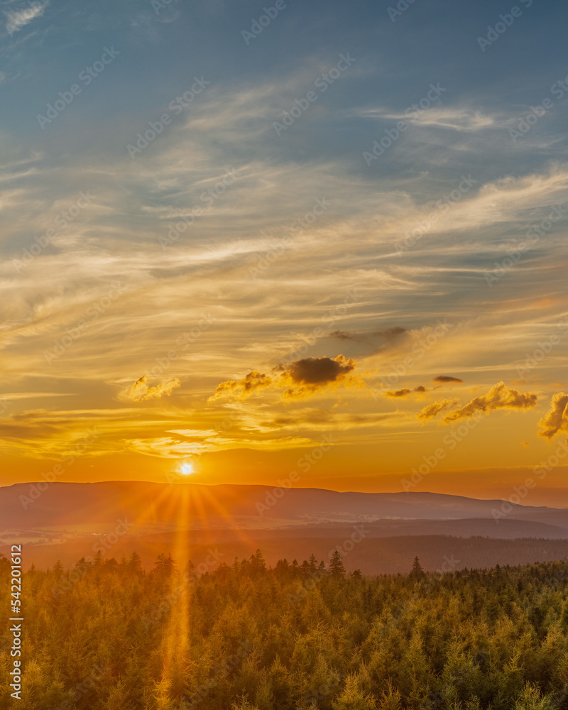 Obraz premium A breathtaking view of a mountain sunset seen from a high vantage point. The warm tones of the setting sun contrast with the cool hues of the rugged mountain landscape, creating a stunning visual disp