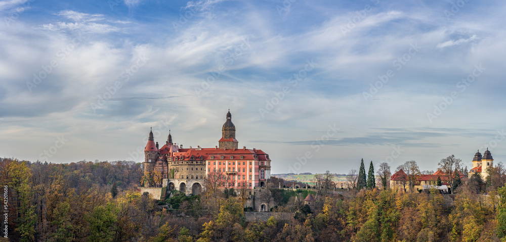 A photo of Książ Castle in Wałbrzych set against a beautiful autumn sky. The castle, with its impressive architecture, stands out against the picturesque background of the colorful fall foliage