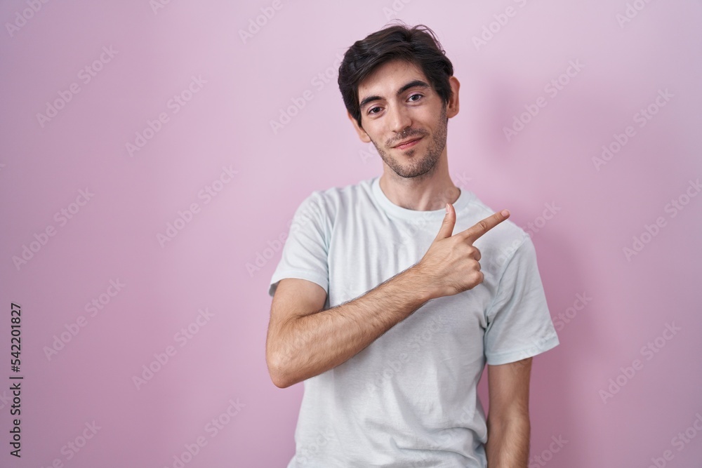 Young hispanic man standing over pink background cheerful with a smile on face pointing with hand and finger up to the side with happy and natural expression Young hispanic man standing over pink background cheerful with a smile on face pointing with hand and finger up to the side with happy and natural expression