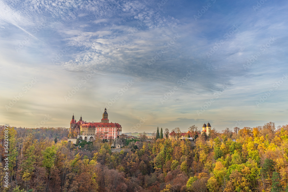 A photo of Książ Castle in Wałbrzych set against a beautiful autumn sky. The castle, with its impressive architecture, stands out against the picturesque background of the colorful fall foliage
