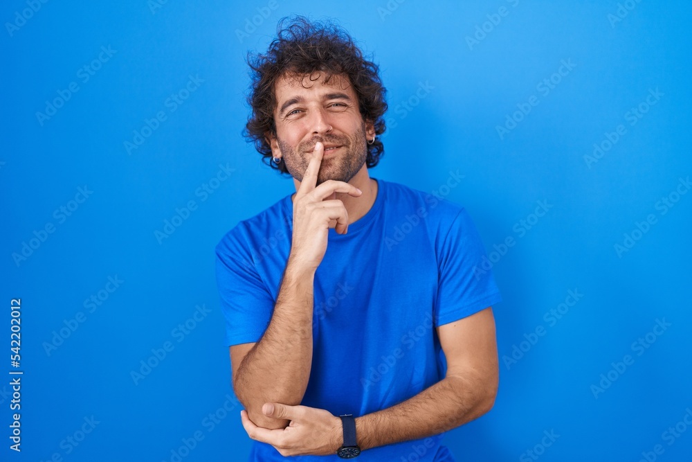 Hispanic young man standing over blue background looking confident at the camera smiling with crossed arms and hand raised on chin. thinking positive. Hispanic young man standing over blue background looking confident at the camera smiling with crossed arms and hand raised on chin. thinking positive.
