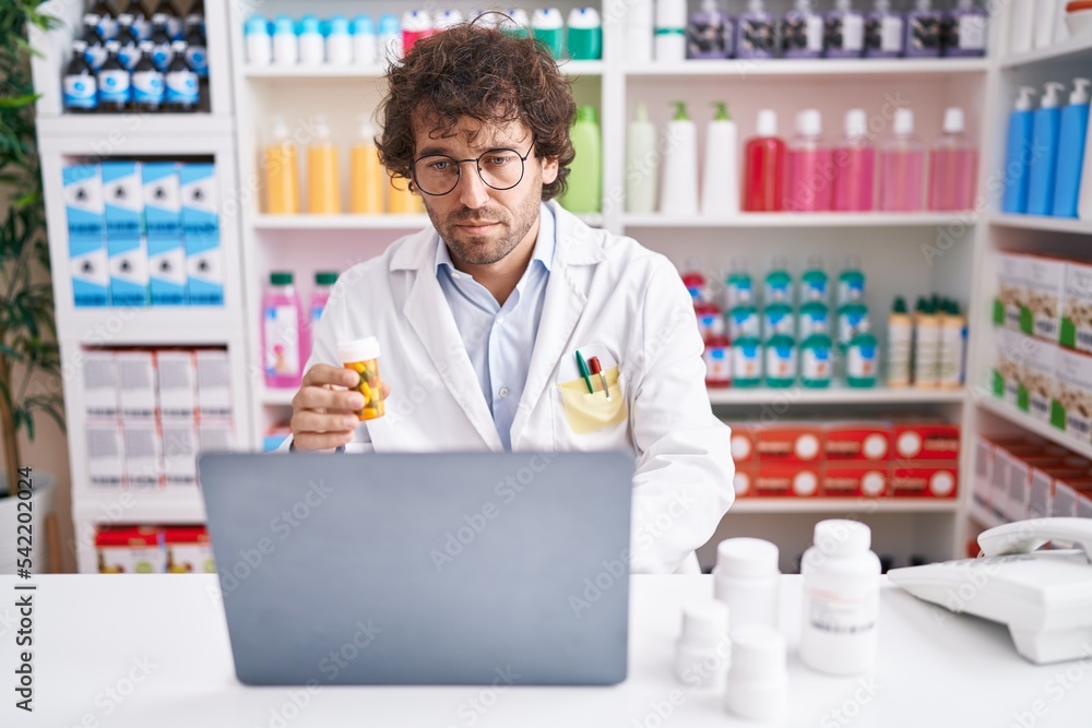 Young hispanic man pharmacist using laptop holding pills bottle at pharmacy Young hispanic man pharmacist using laptop holding pills bottle at pharmacy