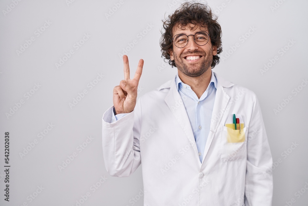Hispanic young man wearing doctor uniform showing and pointing up with fingers number two while smiling confident and happy. Hispanic young man wearing doctor uniform showing and pointing up with fingers number two while smiling confident and happy.