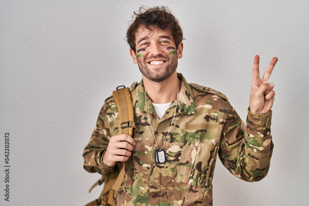 Hispanic young man wearing camouflage army uniform smiling with happy face winking at the camera doing victory sign with fingers. number two. Hispanic young man wearing camouflage army uniform smiling with happy face winking at the camera doing victory sign with fingers. number two.