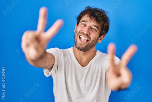 Hispanic young man standing over blue background smiling with tongue out showing fingers of both hands doing victory sign. number two.