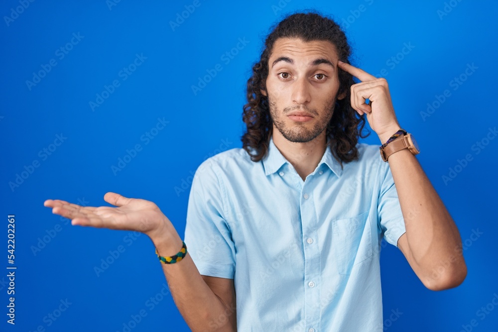Fototapeta premium Young hispanic man standing over blue background confused and annoyed with open palm showing copy space and pointing finger to forehead. think about it.