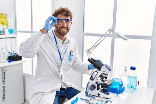 Young hispanic man wearing scientist uniform using microscope at laboratory