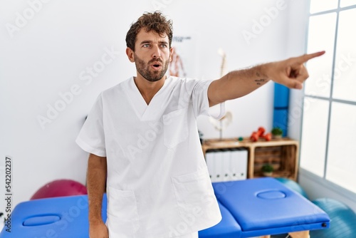 Young handsome physiotherapist man working at pain recovery clinic pointing with finger surprised ahead  open mouth amazed expression  something on the front