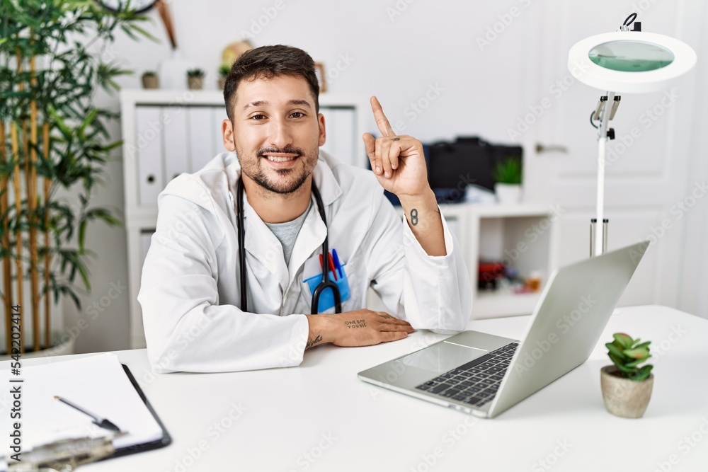 Young doctor working at the clinic using computer laptop showing and pointing up with finger number one while smiling confident and happy.