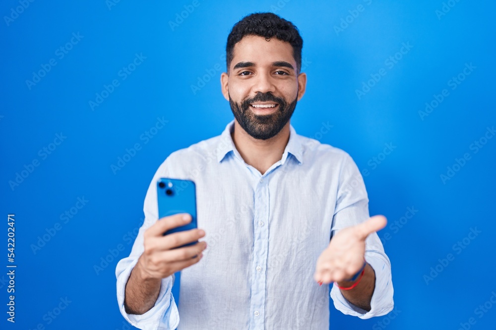 Hispanic man with beard using smartphone typing message smiling cheerful offering palm hand giving assistance and acceptance. Hispanic man with beard using smartphone typing message smiling cheerful offering palm hand giving assistance and acceptance.