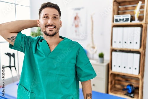 Young physiotherapist man working at pain recovery clinic smiling confident touching hair with hand up gesture, posing attractive and fashionable