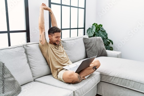 Young hispanic man using laptop stretching arms at home