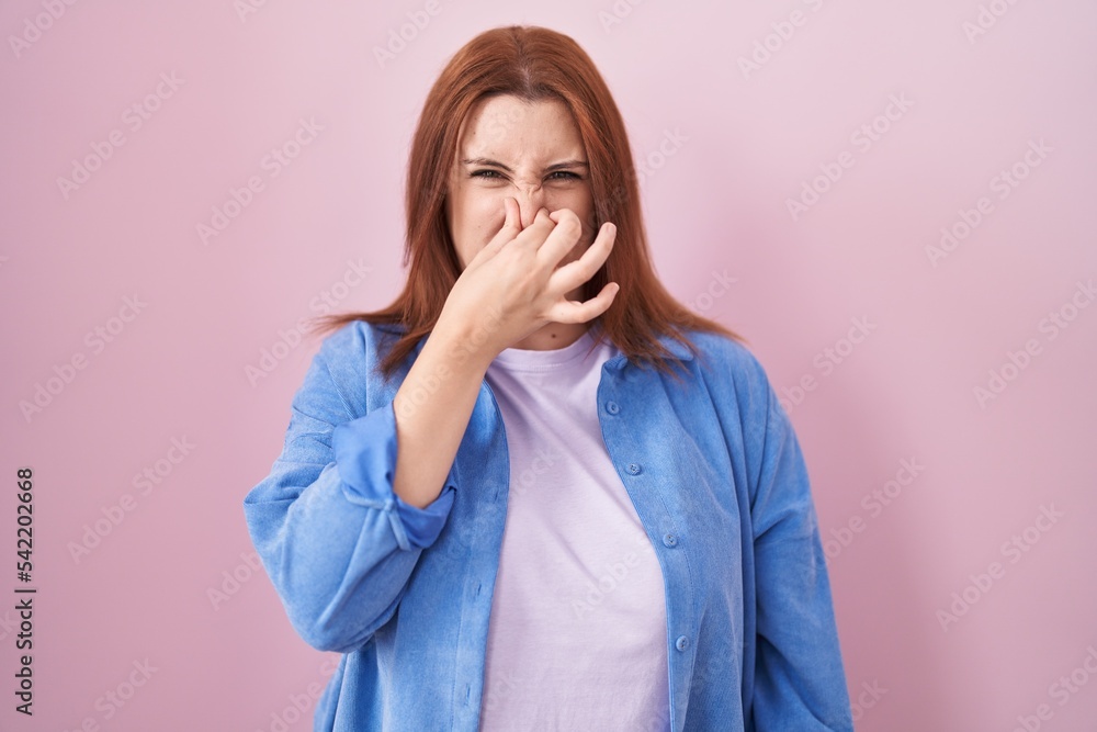 Young hispanic woman with red hair standing over pink background smelling something stinky and disgusting, intolerable smell, holding breath with fingers on nose. bad smell Young hispanic woman with red hair standing over pink background smelling something stinky and disgusting, intolerable smell, holding breath with fingers on nose. bad smell