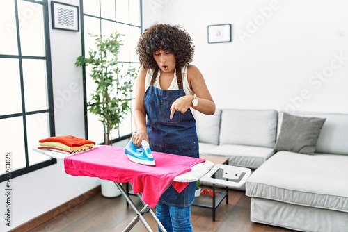 Young middle eastern woman ironing clothes at home pointing down with fingers showing advertisement, surprised face and open mouth