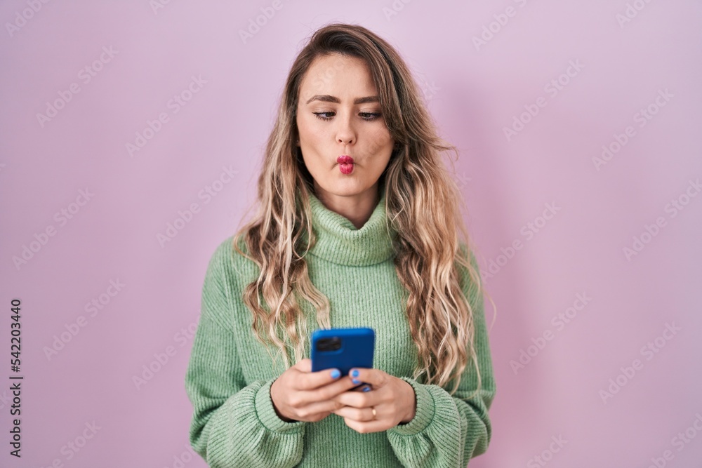 Young caucasian woman using smartphone typing message making fish face with mouth and squinting eyes, crazy and comical. Young caucasian woman using smartphone typing message making fish face with mouth and squinting eyes, crazy and comical.