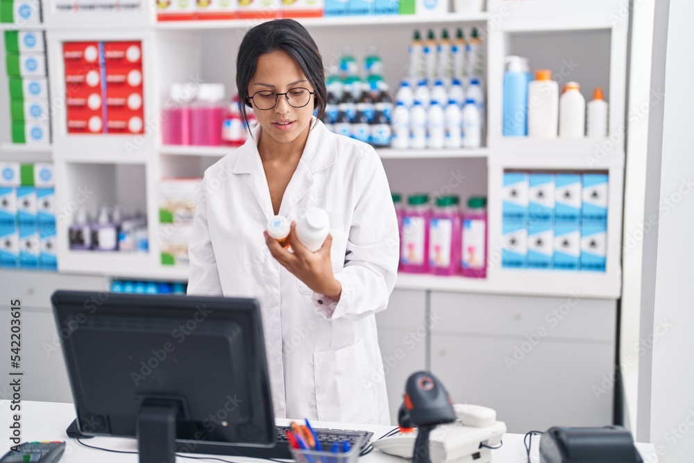 Young hispanic woman pharmacist holding pills bottles using computer at pharmacy Young hispanic woman pharmacist holding pills bottles using computer at pharmacy