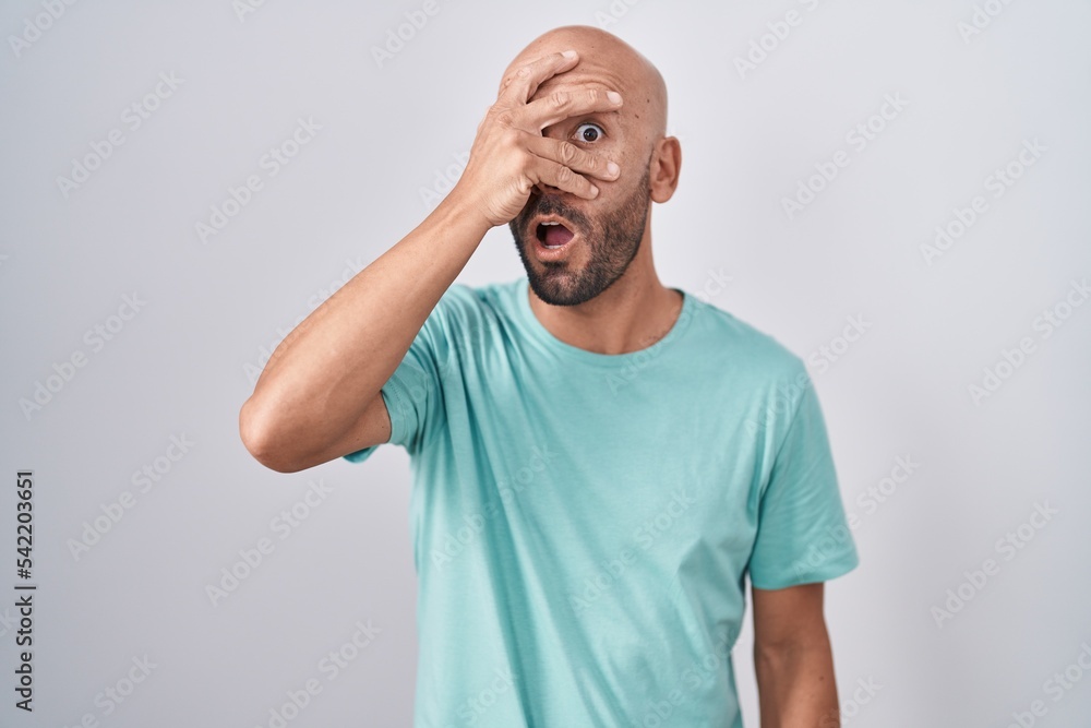 Middle age bald man standing over white background peeking in shock covering face and eyes with hand, looking through fingers with embarrassed expression. Middle age bald man standing over white background peeking in shock covering face and eyes with hand, looking through fingers with embarrassed expression.